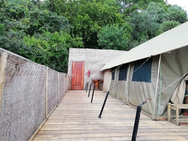 A wooden walkway leads to a canvas tent set up against a lush, green forest backdrop. The tent has several windows and is connected to a woven bamboo wall on one side. A wooden door and a fire extinguisher are visible at the end of the walkway, adding elements of utility and safety.