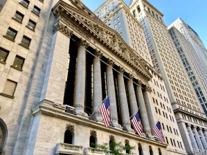 A grand neoclassical building with a row of tall columns and ornate detailing. American flags are prominently displayed, hanging from the facade. Surrounding the structure are other tall buildings under a clear blue sky.