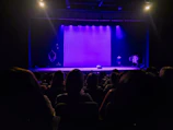 A dimly lit theater with an audience seated facing a stage. The stage features a purple backdrop and various props. The lighting creates a dramatic and anticipatory atmosphere.