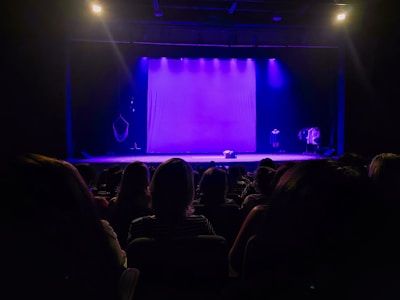 A dimly lit theater with an audience seated facing a stage. The stage features a purple backdrop and various props. The lighting creates a dramatic and anticipatory atmosphere.