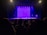 A dimly lit theater with an audience seated facing a stage. The stage features a purple backdrop and various props. The lighting creates a dramatic and anticipatory atmosphere.