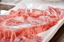 A close-up of raw, marbled beef slices arranged on a rectangular white plate. The meat is rich in color with visible patterns of intramuscular fat, indicating high-quality cuts.