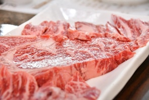A close-up of raw, marbled beef slices arranged on a rectangular white plate. The meat is rich in color with visible patterns of intramuscular fat, indicating high-quality cuts.