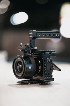 A compact digital camera housed in a black metal cage with mounting options. The camera lens is prominently labeled, and the overall setup rests on a flat surface. The background is softly blurred, highlighting the camera's details.