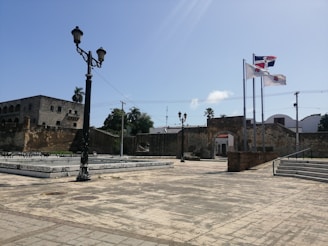 A historic plaza with stone-paved ground, featuring a tall black street lamp in the foreground. In the background, there's an old stone building with arched windows and a wall with a small doorway. Three flagpoles display national and regional flags, waving in the breeze. Lush greenery, including palm trees, is visible against a clear blue sky.