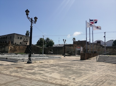 A historic plaza with stone-paved ground, featuring a tall black street lamp in the foreground. In the background, there's an old stone building with arched windows and a wall with a small doorway. Three flagpoles display national and regional flags, waving in the breeze. Lush greenery, including palm trees, is visible against a clear blue sky.