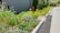 A well-maintained garden with a variety of colorful flowers and lush green plants lines a paved path. A sign with an accessibility symbol is visible. The environment exudes a sense of tidiness and natural beauty.