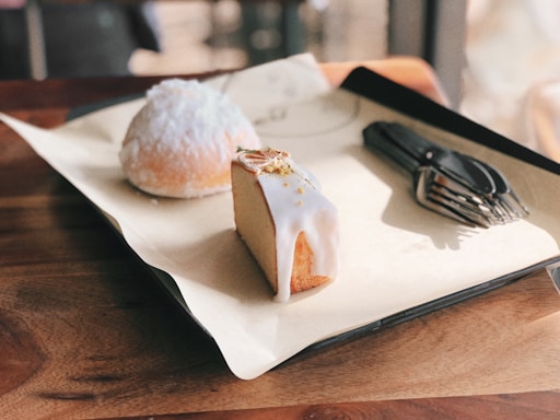 A slice of sponge cake with white icing sits on a tray alongside a round pastry dusted with powdered sugar. Several black plastic forks are placed next to them. They are arranged on a wooden table, reflecting a warm, inviting ambiance.