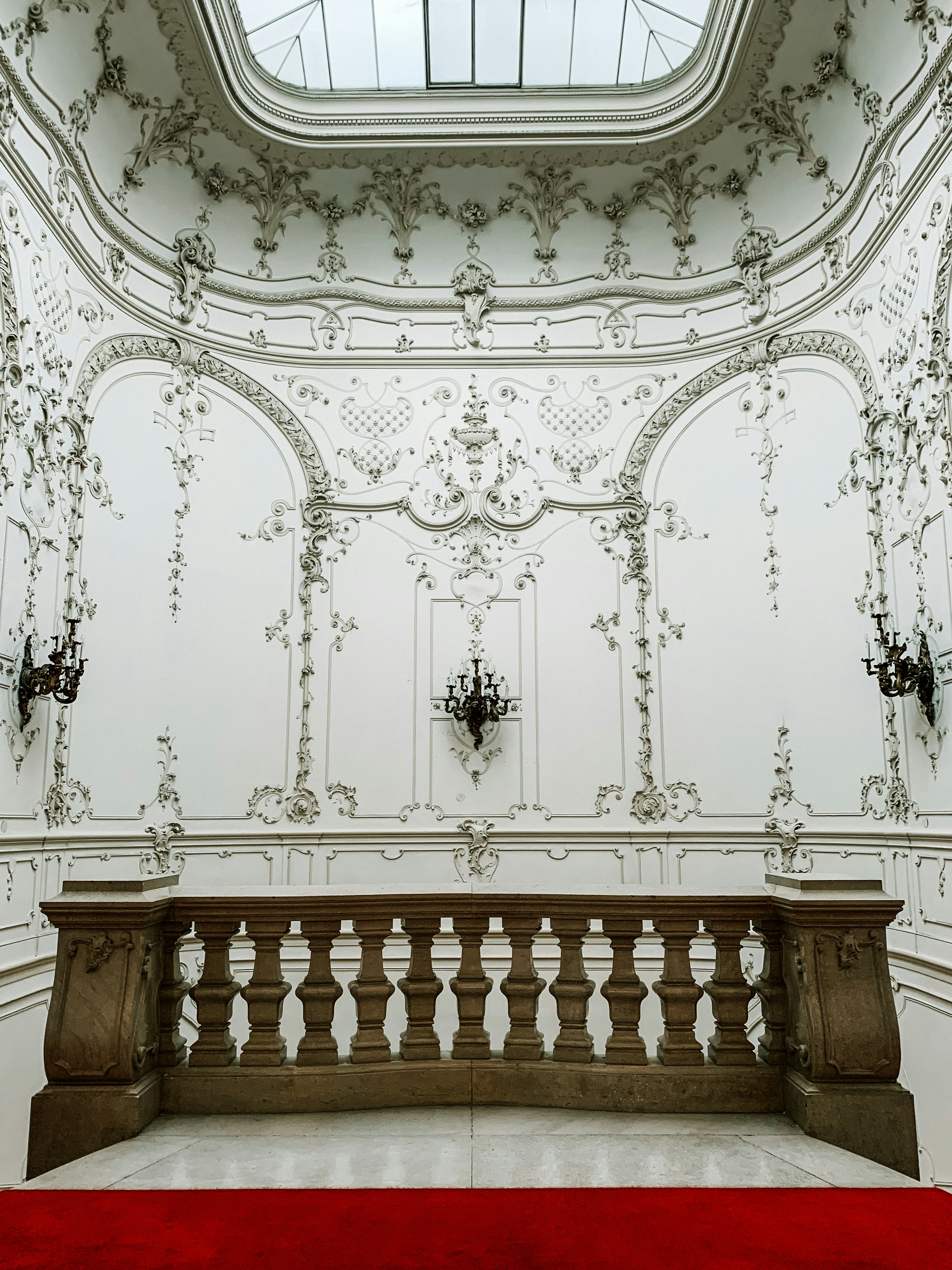 An opulent and intricately designed interior space featuring ornate plasterwork and moldings on the walls. A stone balustrade separates a lower area covered by a red carpet. Above, a skylight allows natural light to illuminate the space, highlighting the elaborate floral and scroll patterns.