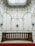An opulent and intricately designed interior space featuring ornate plasterwork and moldings on the walls. A stone balustrade separates a lower area covered by a red carpet. Above, a skylight allows natural light to illuminate the space, highlighting the elaborate floral and scroll patterns.