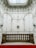 An opulent and intricately designed interior space featuring ornate plasterwork and moldings on the walls. A stone balustrade separates a lower area covered by a red carpet. Above, a skylight allows natural light to illuminate the space, highlighting the elaborate floral and scroll patterns.