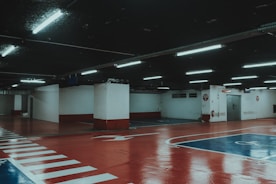 A dimly lit underground parking garage with a glossy red and blue painted floor and white markings. Fluorescent lights hang from the ceiling. The columns and walls are painted in a white and red scheme.