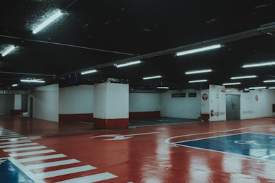 A dimly lit underground parking garage with a glossy red and blue painted floor and white markings. Fluorescent lights hang from the ceiling. The columns and walls are painted in a white and red scheme.
