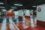 An underground parking lot with a polished, reflective floor painted in red and blue hues. Bright fluorescent lights illuminate the space, casting reflections on the glossy surface. Concrete pillars support the structure, with one prominently displaying the number '2330 V' in white against a red circle.