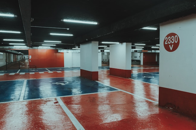 An underground parking lot with a polished, reflective floor painted in red and blue hues. Bright fluorescent lights illuminate the space, casting reflections on the glossy surface. Concrete pillars support the structure, with one prominently displaying the number '2330 V' in white against a red circle.