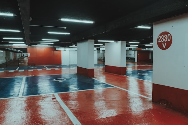 An underground parking lot with a polished, reflective floor painted in red and blue hues. Bright fluorescent lights illuminate the space, casting reflections on the glossy surface. Concrete pillars support the structure, with one prominently displaying the number '2330 V' in white against a red circle.