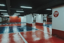 An underground parking lot with a polished, reflective floor painted in red and blue hues. Bright fluorescent lights illuminate the space, casting reflections on the glossy surface. Concrete pillars support the structure, with one prominently displaying the number '2330 V' in white against a red circle.