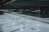 An empty, well-lit indoor parking garage with marked parking spaces and overhead fluorescent lights. The floor is polished and reflects the artificial lighting, and there are signs and directional arrows painted on the ground.