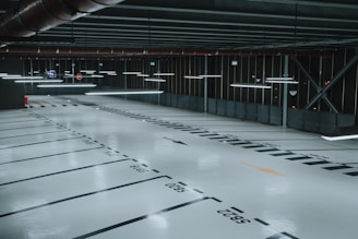 An empty, well-lit indoor parking garage with marked parking spaces and overhead fluorescent lights. The floor is polished and reflects the artificial lighting, and there are signs and directional arrows painted on the ground.