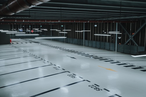 An empty, well-lit indoor parking garage with marked parking spaces and overhead fluorescent lights. The floor is polished and reflects the artificial lighting, and there are signs and directional arrows painted on the ground.