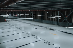 An empty, well-lit indoor parking garage with marked parking spaces and overhead fluorescent lights. The floor is polished and reflects the artificial lighting, and there are signs and directional arrows painted on the ground.