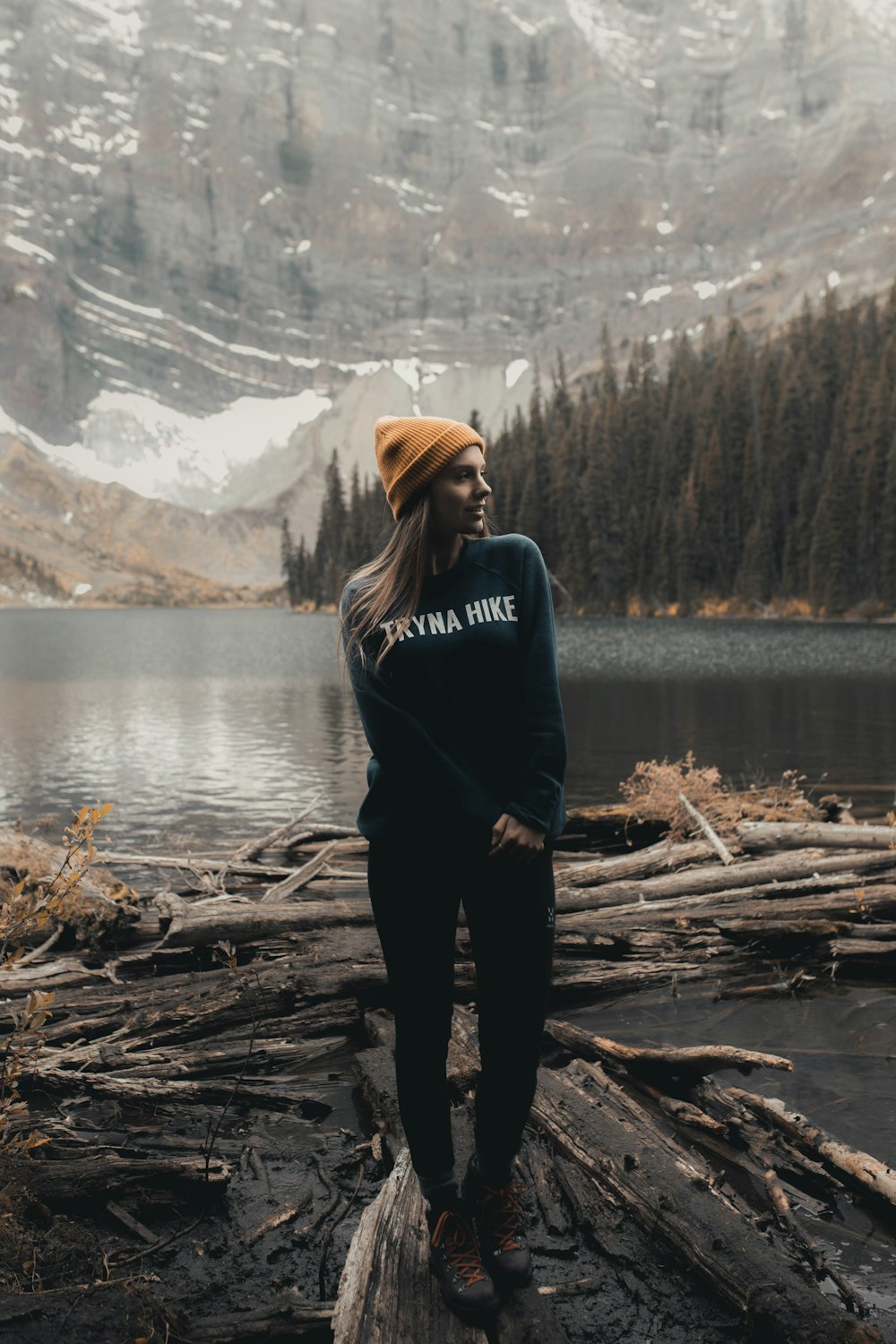 Donna in felpa con cappuccio nera in piedi su tronco di legno marrone vicino al lago durante il giorno