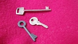 Three different keys are placed on a textured pink surface. One key is long and metallic, while another is circular with a grooved design. The third key is more traditional, with ridges and a rounded handle.
