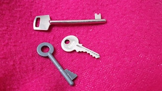 Three different keys are placed on a textured pink surface. One key is long and metallic, while another is circular with a grooved design. The third key is more traditional, with ridges and a rounded handle.