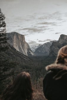 A breathtaking view of a vast valley surrounded by majestic cliffs and forested mountains. Snow-capped peaks are visible in the distance. The foreground includes silhouettes of two people admiring the natural landscape, adding a sense of scale and appreciation.