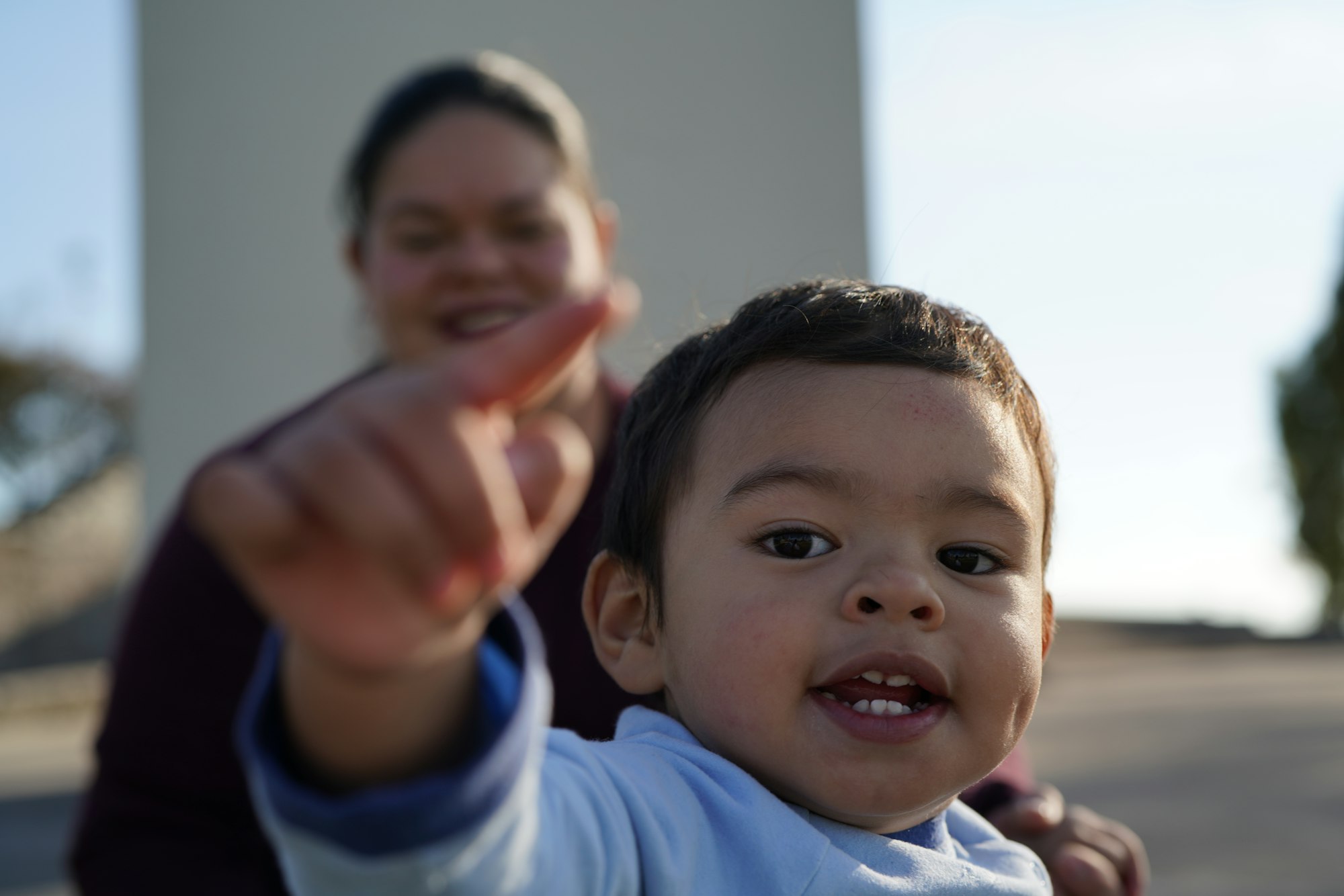 A baby points and smiles at the camera, while his mother looks on from the background.