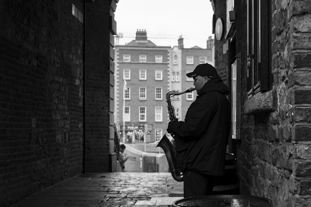 man in black jacket playing saxophone sitting on concrete bench in grayscale photography