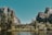 A serene natural landscape featuring towering rock formations partially covered with vegetation, a flowing waterfall cascading down the rock face, and a lush forest in the foreground. A calm river flows gently at the bottom, reflecting the clear blue sky above.