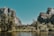 A serene natural landscape featuring towering rock formations partially covered with vegetation, a flowing waterfall cascading down the rock face, and a lush forest in the foreground. A calm river flows gently at the bottom, reflecting the clear blue sky above.