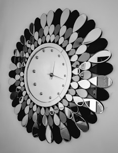 A large decorative wall clock with a circular face surrounded by petal-shaped mirror pieces arranged in a radial pattern. The clock has a simple design with a minimalistic face, small hour markers, and two hands indicating the time. The background is a plain wall, emphasizing the clock's intricate design.