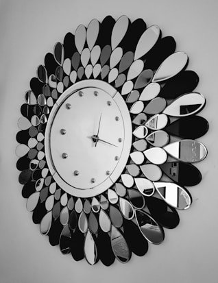 A large decorative wall clock with a circular face surrounded by petal-shaped mirror pieces arranged in a radial pattern. The clock has a simple design with a minimalistic face, small hour markers, and two hands indicating the time. The background is a plain wall, emphasizing the clock's intricate design.