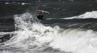 A surfer is performing an aerial maneuver above a large, breaking wave in turbulent ocean waters. The surfer, who is upside down, is gripping the surfboard tightly while navigating through the challenging waves.