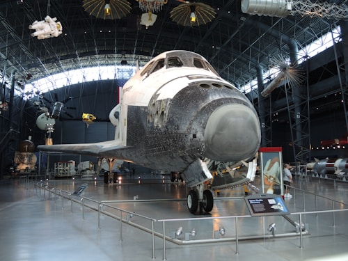 A space shuttle is exhibited inside a large museum hangar. The shuttle is displayed on the ground with its nose facing forward. Various other aerospace artifacts and equipment are suspended from the ceiling and positioned throughout the room. The hangar features a high arched ceiling made of metal and glass, allowing natural light to illuminate the display area.
