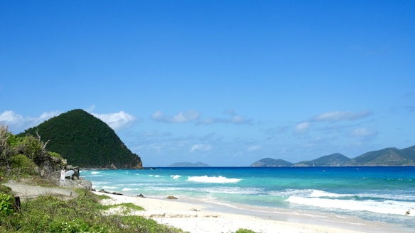 A serene beach landscape with clear blue skies and lush green hills in the background. The turquoise ocean waves gently lap the sandy shoreline, while a small path with vegetation leads down to the beach.