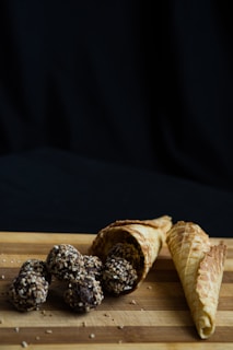 A collection of chocolate truffles coated with chopped nuts is placed on a wooden surface. Next to the truffles, there are waffle cones empty and leaning on their side. The background is plain and dark, creating a contrast with the light-colored cones and wooden surface.