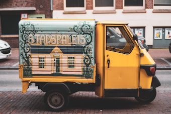 A small, yellow utility vehicle is parked on a street. The vehicle features an artistic side panel with a mural depicting a building labeled 'STADSPALEIS'. The background shows a brick wall with windows and posters.