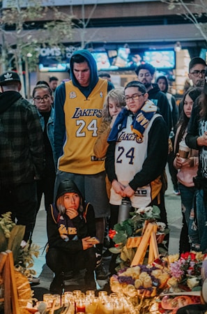 A group of people gather around a memorial with lit candles and flowers. Several individuals are wearing Lakers jerseys with the number 24, reflecting a somber and respectful atmosphere.