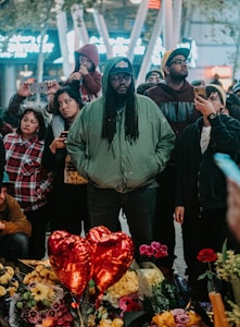 A diverse group of people gathered in an outdoor setting during the evening, some holding or pointing their phones. At the forefront, a man with long hair and a green jacket stands solemnly. In front of them are red heart-shaped balloons, various flowers, and bouquets. The atmosphere suggests a memorial or vigil.