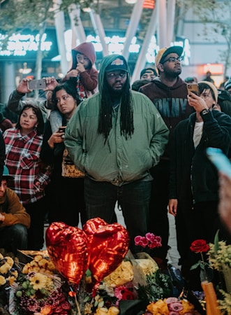 A diverse group of people gathered in an outdoor setting during the evening, some holding or pointing their phones. At the forefront, a man with long hair and a green jacket stands solemnly. In front of them are red heart-shaped balloons, various flowers, and bouquets. The atmosphere suggests a memorial or vigil.