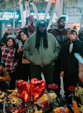 A diverse group of people gathered in an outdoor setting during the evening, some holding or pointing their phones. At the forefront, a man with long hair and a green jacket stands solemnly. In front of them are red heart-shaped balloons, various flowers, and bouquets. The atmosphere suggests a memorial or vigil.
