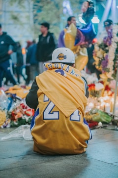 A person is kneeling in front of a memorial, wearing a yellow basketball jersey with the number 24. The scene is surrounded by flowers, candles, and blurred figures of people in the background, creating a somber atmosphere.
