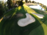 An aerial view of a golf course featuring neatly maintained greens. Sand bunkers are prominent in the center, surrounded by lush grass. Shadows from tall trees stretch across the ground, with a calm body of water visible in the distance. The sunlight casts a warm glow across the landscape, highlighting the contours of the course.