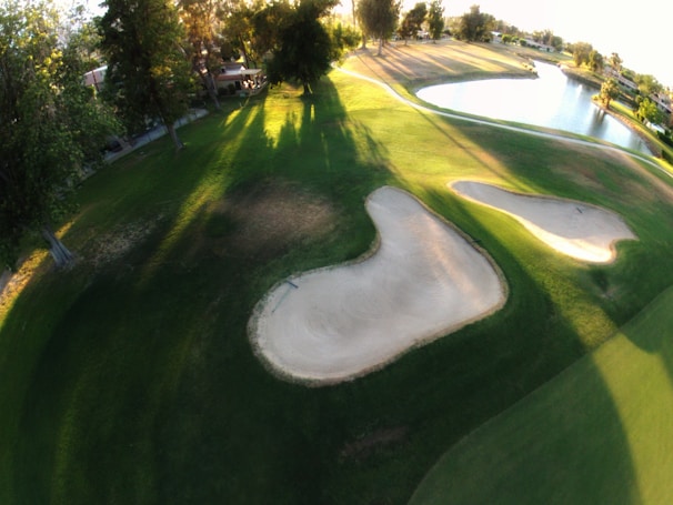 An aerial view of a golf course featuring neatly maintained greens. Sand bunkers are prominent in the center, surrounded by lush grass. Shadows from tall trees stretch across the ground, with a calm body of water visible in the distance. The sunlight casts a warm glow across the landscape, highlighting the contours of the course.