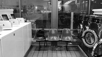 Interior of a laundromat with rows of washing machines and dryers on either side. In the center, there are two plastic chairs with a black bag placed on one. The space is lit with fluorescent lighting, and large windows provide a view of the outside, showing reflections and faint outlines of another room or building.