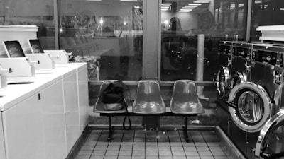 Interior of a laundromat with rows of washing machines and dryers on either side. In the center, there are two plastic chairs with a black bag placed on one. The space is lit with fluorescent lighting, and large windows provide a view of the outside, showing reflections and faint outlines of another room or building.