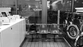 Interior of a laundromat with rows of washing machines and dryers on either side. In the center, there are two plastic chairs with a black bag placed on one. The space is lit with fluorescent lighting, and large windows provide a view of the outside, showing reflections and faint outlines of another room or building.
